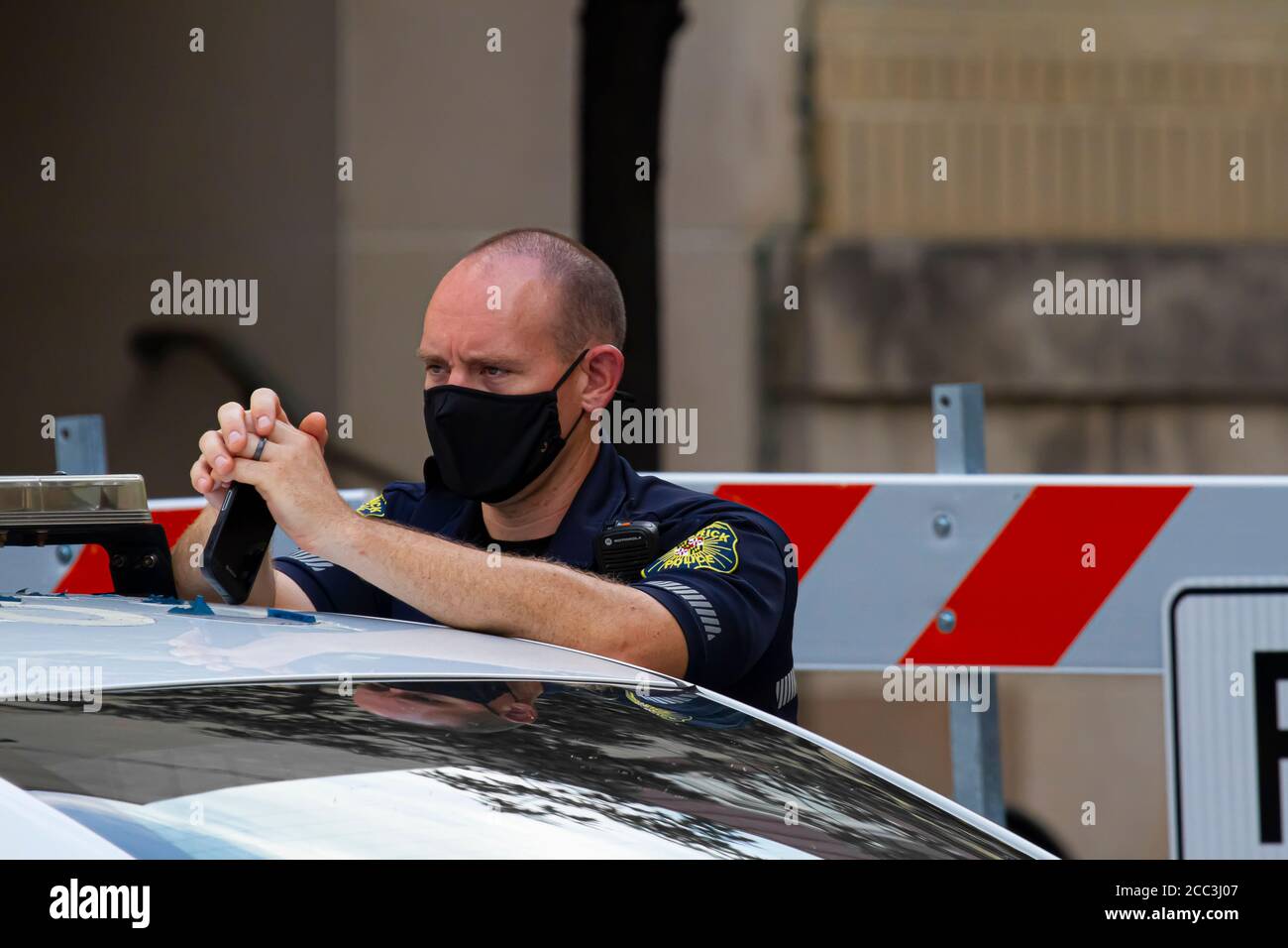 Frederick, MD, USA 08/14/2020: Un giovane poliziotto maschio caucasico indossa una maschera mentre è in servizio. È in piedi presso la sua macchina di polizia di fronte o Foto Stock