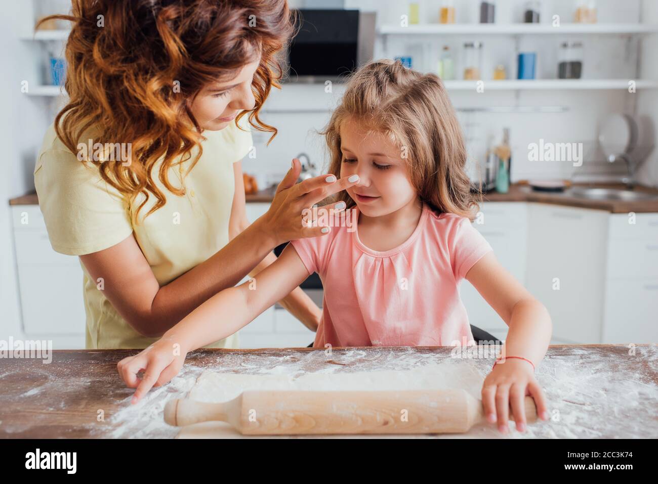 madre che tocca il naso della figlia che rotola fuori l'impasto sul tavolo sparso con farina Foto Stock