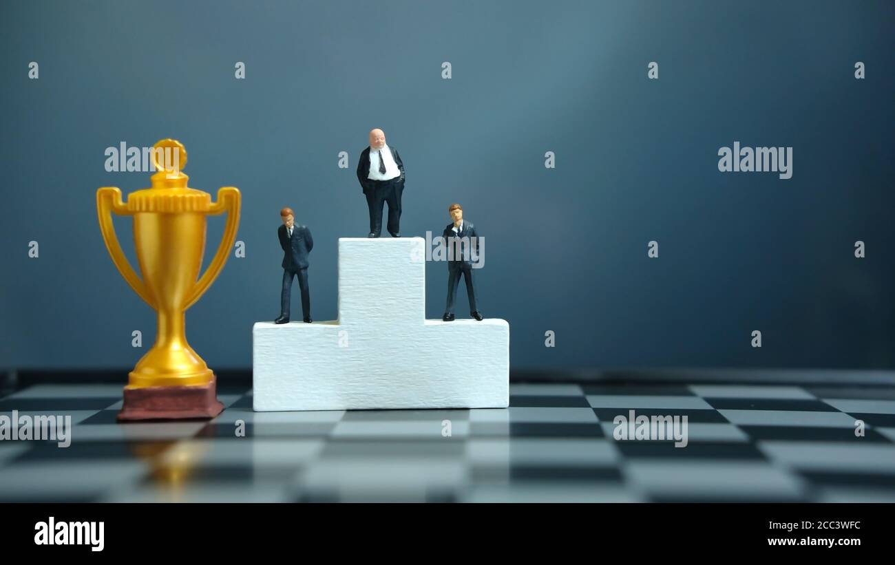 Concetto di business in miniatura - uomo d'affari in piedi sul podio bianco vincitore con trofeo d'oro e d'argento Foto Stock
