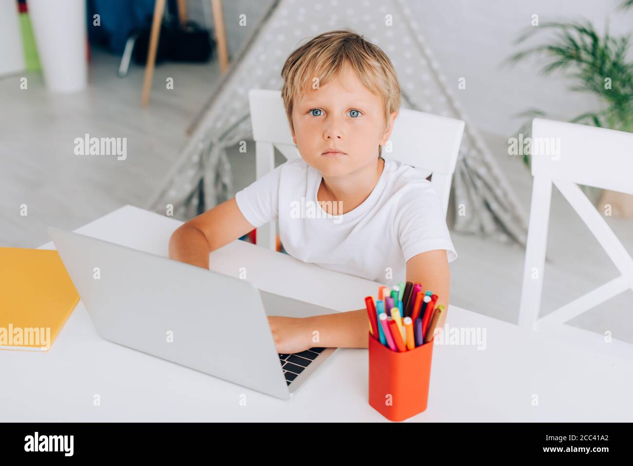 vista ad alto angolo del ragazzo in una t-shirt bianca con un computer portatile mentre si siede al tavolo vicino porta penna e bambini wigwam sullo sfondo Foto Stock