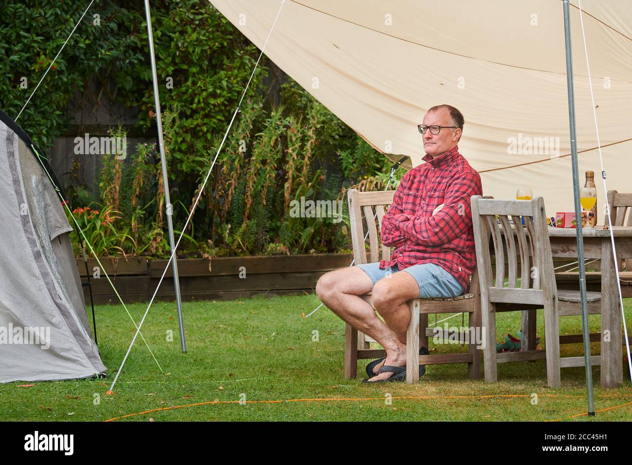 Un uomo di mezza età si siede armato di croce nel pensiero pensivo sotto una tenda awnng in un giorno piovoso nella sua vacanza di giardino posteriore staycation. Foto Stock