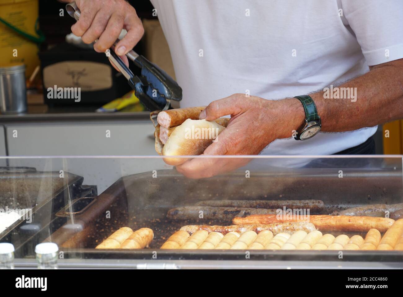 Un uomo che prepara e una salsiccia alla griglia in un pane cuocia con cipolla tostata per un cliente in un veloce Cabina di ristorazione in un mercato di Friburgo am Breisgau Foto Stock