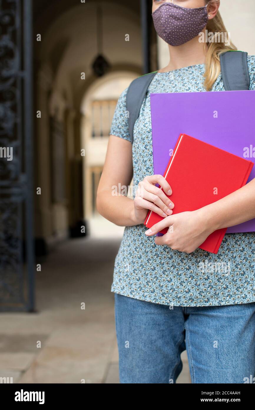 Primo piano di una studentessa femminile che si trova al di fuori del college o dell'università Costruzione indossare maschera viso durante Covid-19 Pandemic Foto Stock