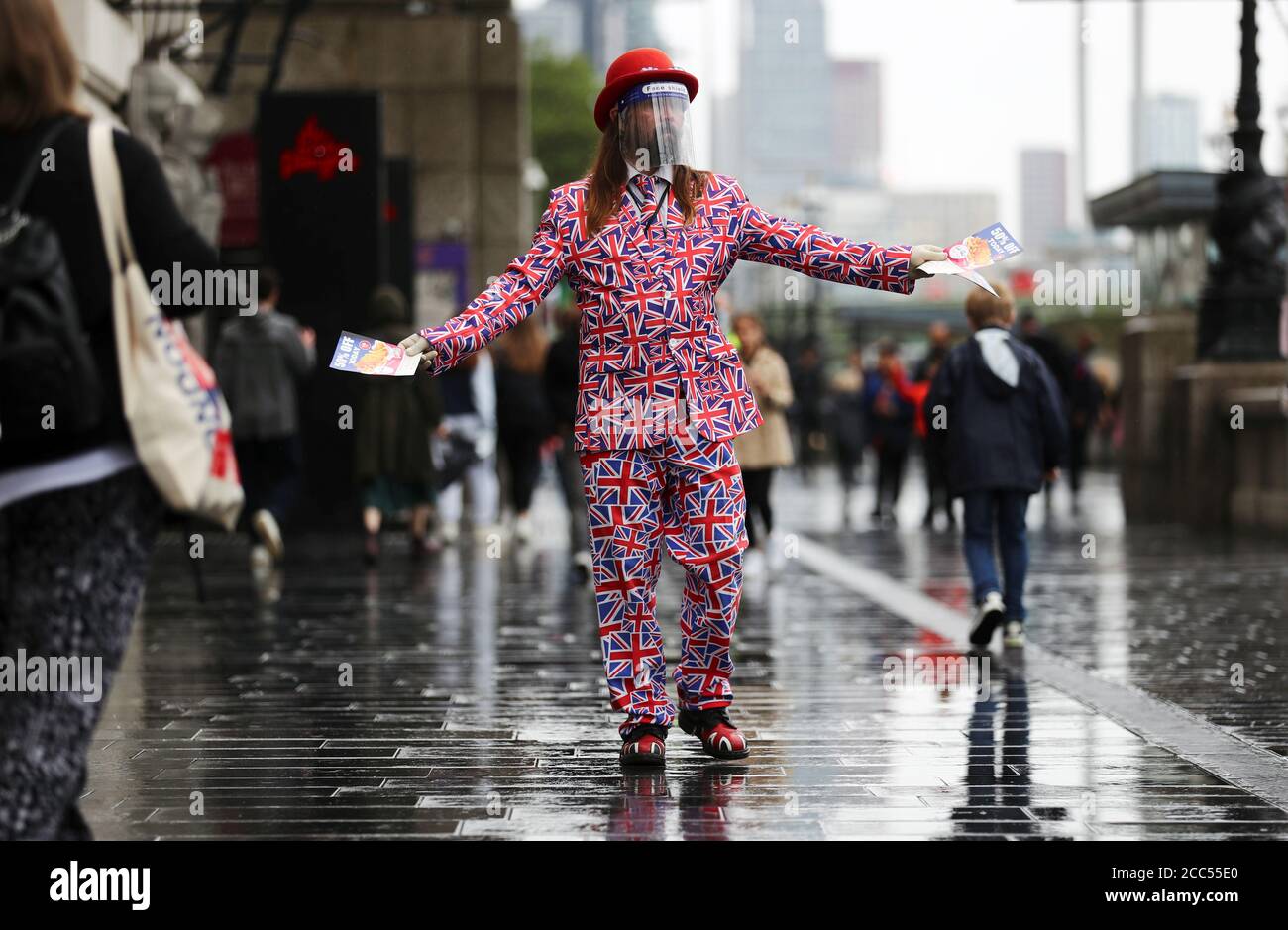 Un uomo in una tuta bandiera Union Jack consegna volantini a persone sotto la pioggia alla Queen's Walk di Londra, Gran Bretagna 19 agosto 2020. REUTERS/Simon Dawson Foto Stock
