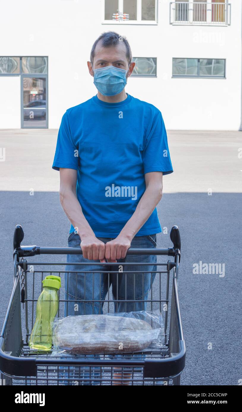 Uomo che indossa una maschera medica in piedi con un carrello quasi vuoto di fronte a un negozio durante la pandemia di coronavirus. Concetto di crisi economica Ala Foto Stock
