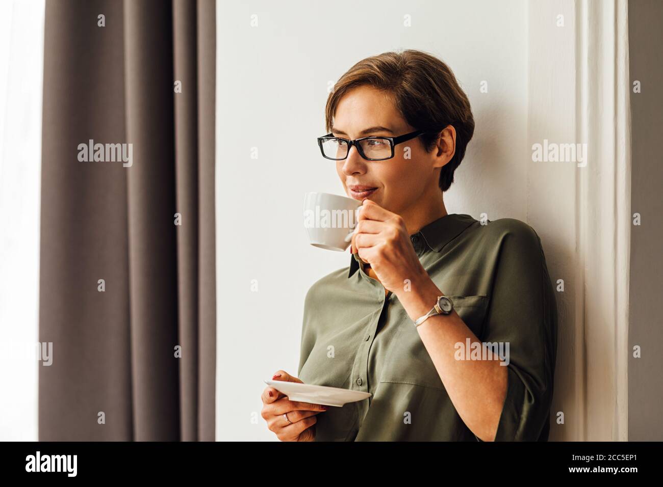 Donna mid adult in abiti formali con caffè in camera d'albergo e guardando via. Donna d'affari in piedi a muro in appartamento e bere caffè. Foto Stock