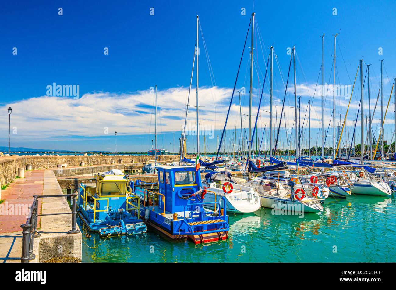 Desenzano del Garda, Italia, 11 settembre 2019: Molo in pietra con yacht sul posto barca porto di parcheggio e faro, blu cielo bianco nuvole sfondo, Lombardia Foto Stock