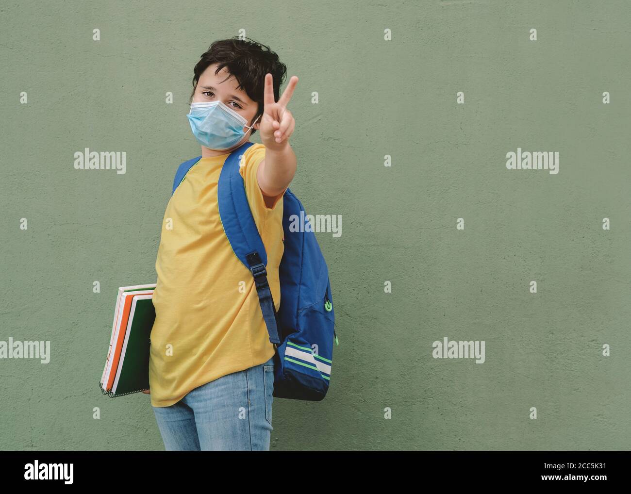 covid-19, capretto con maschera medica e zaino andando a scuola fare segno di vittoria su sfondo verde Foto Stock
