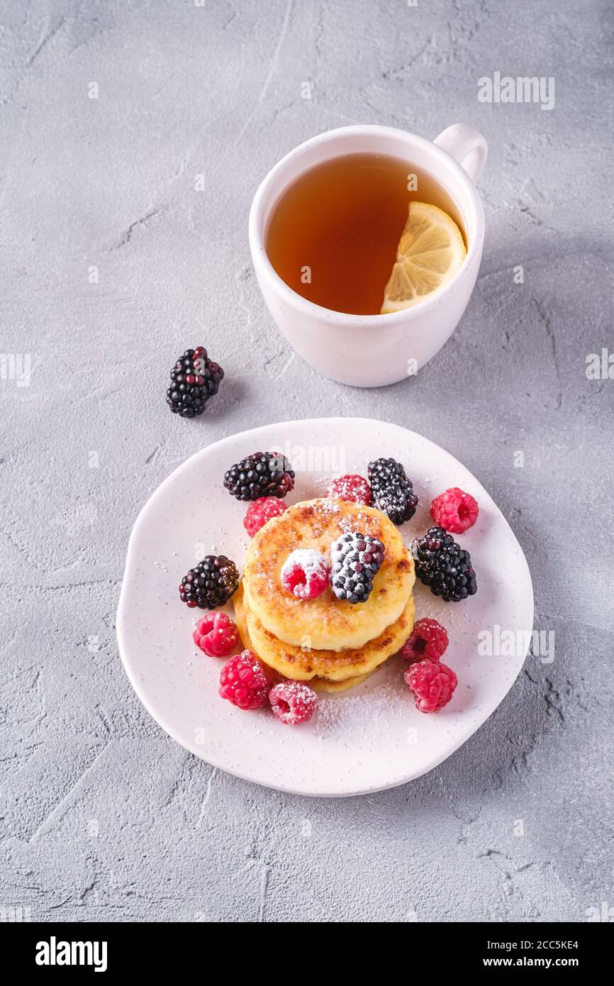 Frittelle al formaggio e zucchero in polvere, frittelle di cagliata dessert con lampone e frutti di bosco in piatto vicino alla tazza di tè caldo con fetta di limone Foto Stock
