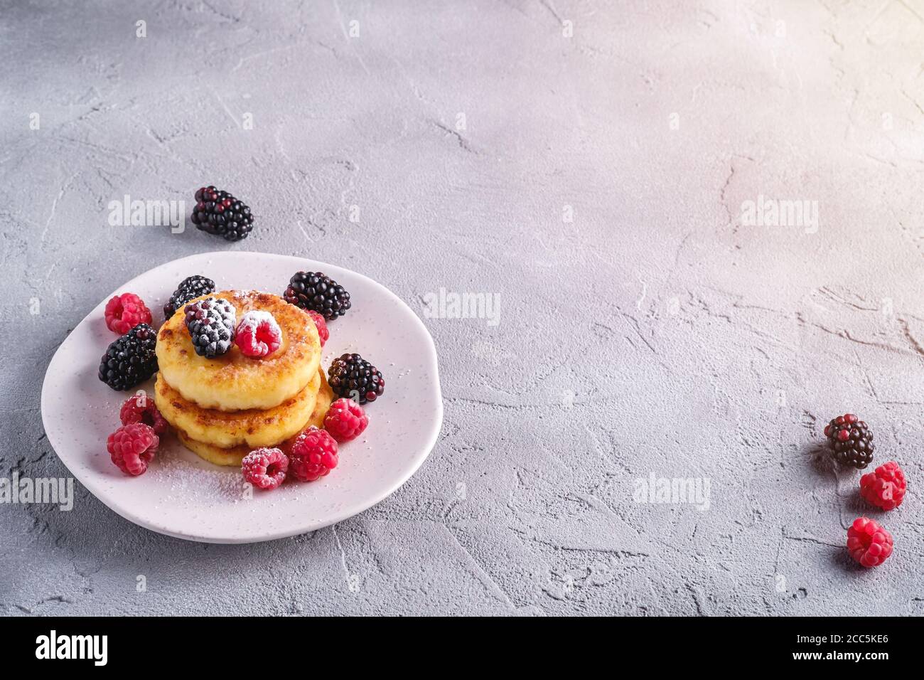 Frittelle al formaggio di cottage e zucchero in polvere, frittelle di cagliata dessert con lampone e bacche di mora in piastra su fondo di cemento di pietra Foto Stock