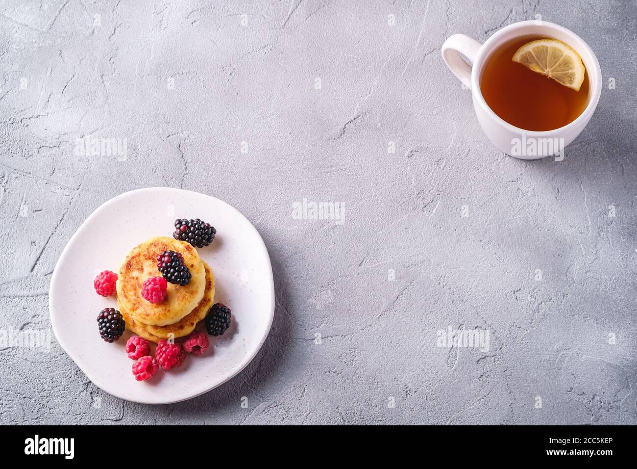 Frittelle di formaggio cottage, frittelle di cagliata dessert con lampone e bacche di mora in piatto vicino alla tazza di tè caldo con fetta di limone su cemento di pietra Foto Stock