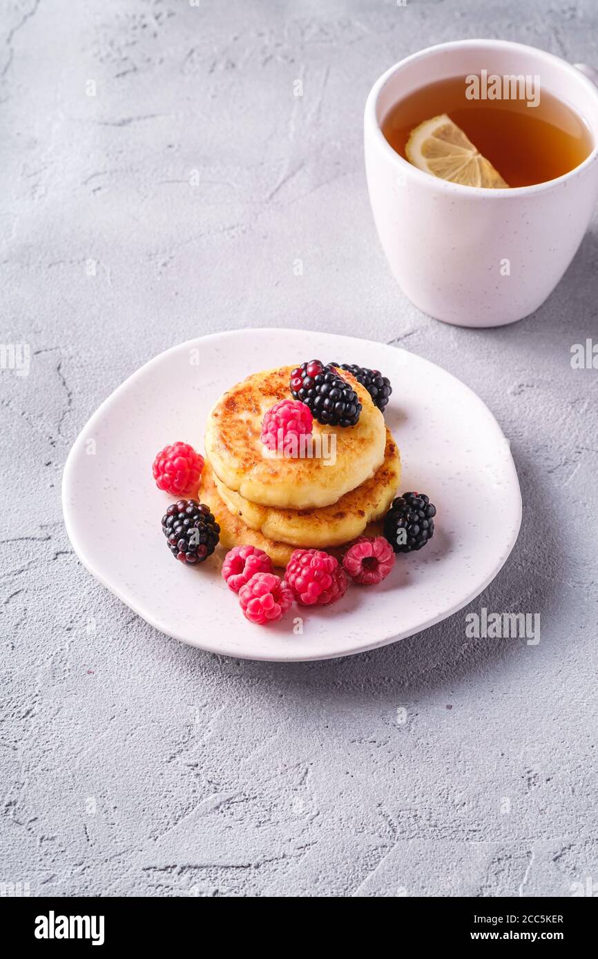 Frittelle di formaggio cottage, frittelle di cagliata dessert con lampone e bacche di mora in piatto vicino alla tazza di tè caldo con fetta di limone su cemento di pietra Foto Stock
