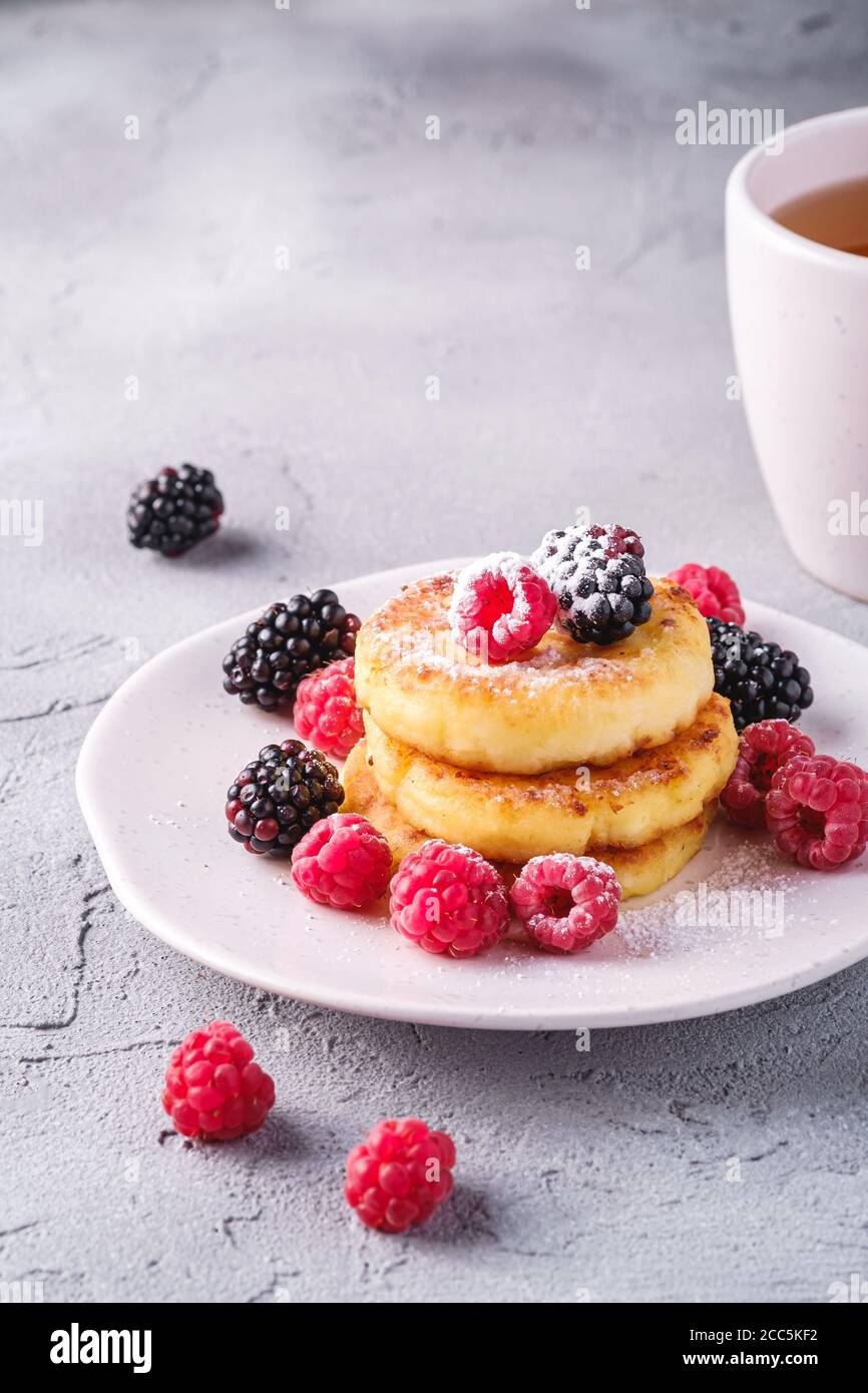 Frittelle al formaggio e zucchero in polvere, frittelle di cagliata dessert con lampone e frutti di bosco in piatto vicino alla tazza di tè caldo con fetta di limone Foto Stock