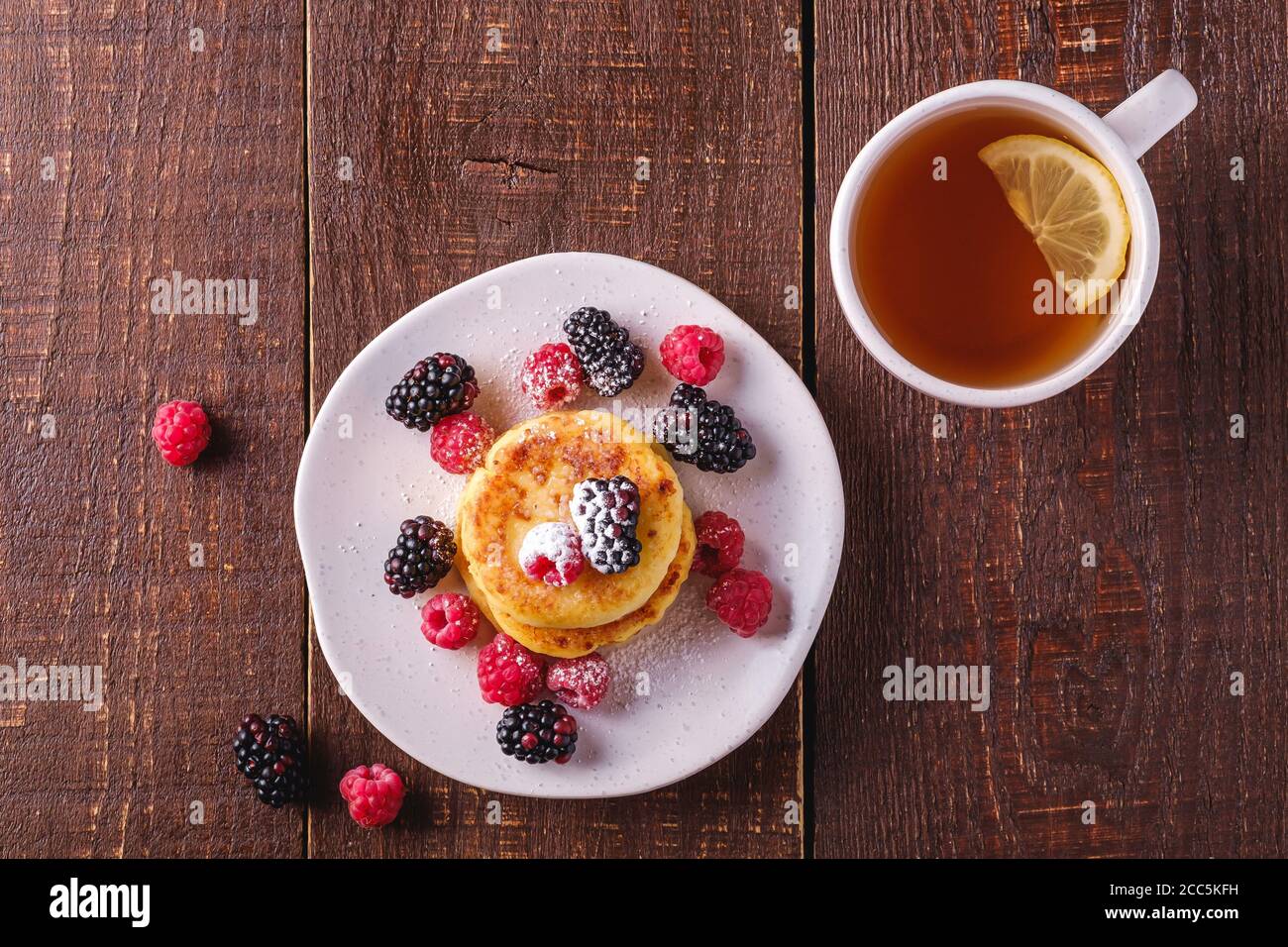Frittelle di formaggio cottage, frittelle di cagliata dessert con lampone e frutti di bosco in piatto vicino alla tazza di tè caldo con fetta di limone Foto Stock