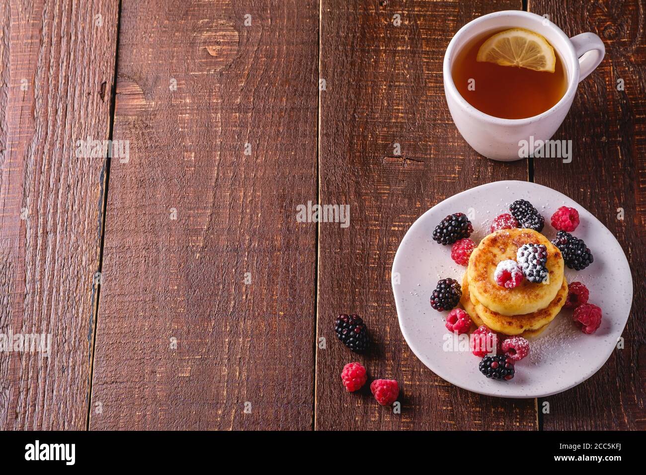 Frittelle di formaggio cottage, frittelle di cagliata dessert con lampone e bacche di mora in piatto vicino alla tazza di tè caldo con fetta di limone su legno marrone scuro Foto Stock