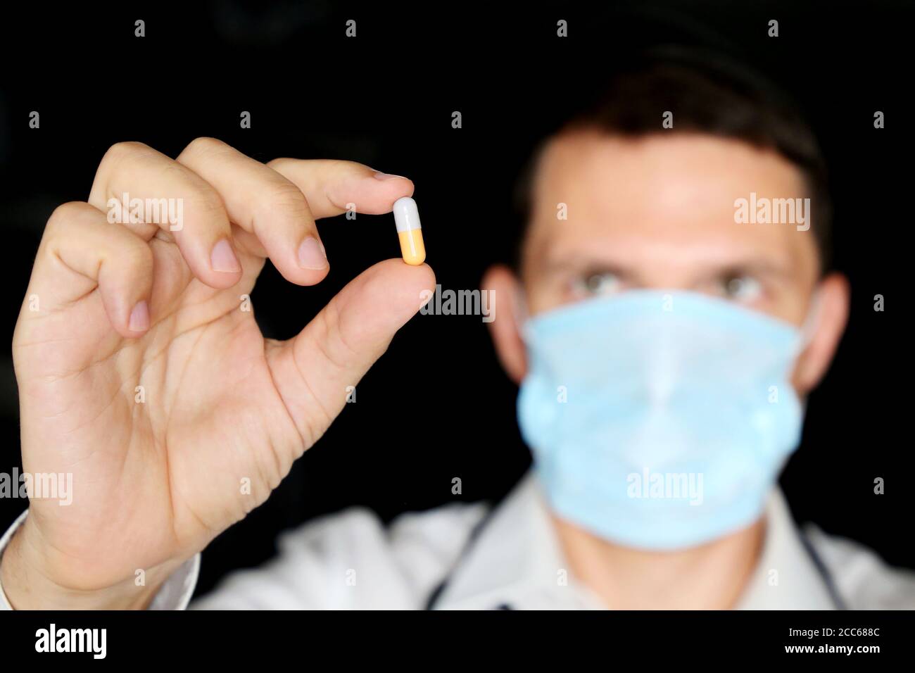 Medico in maschera medica tenendo pillola, mano maschile con farmaco in capsula primo piano. Concetto di farmacista, farmaci, antibiotici o vitamine Foto Stock