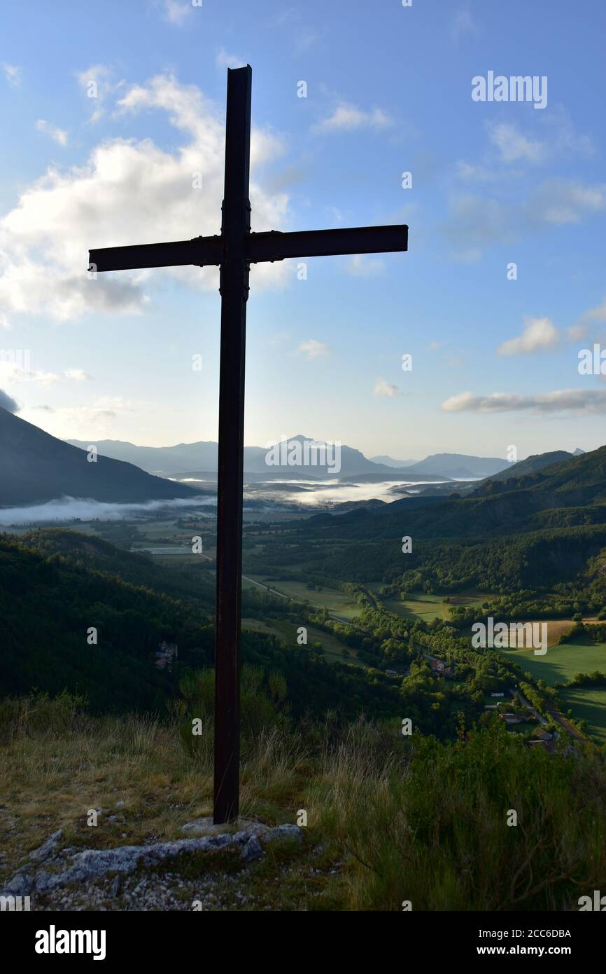 Croce cristiana su una montagna, mattina presto, cielo blu, nebbia nella valle, montagne sullo sfondo, la Beaume, Hautes Alpes, Francia Foto Stock