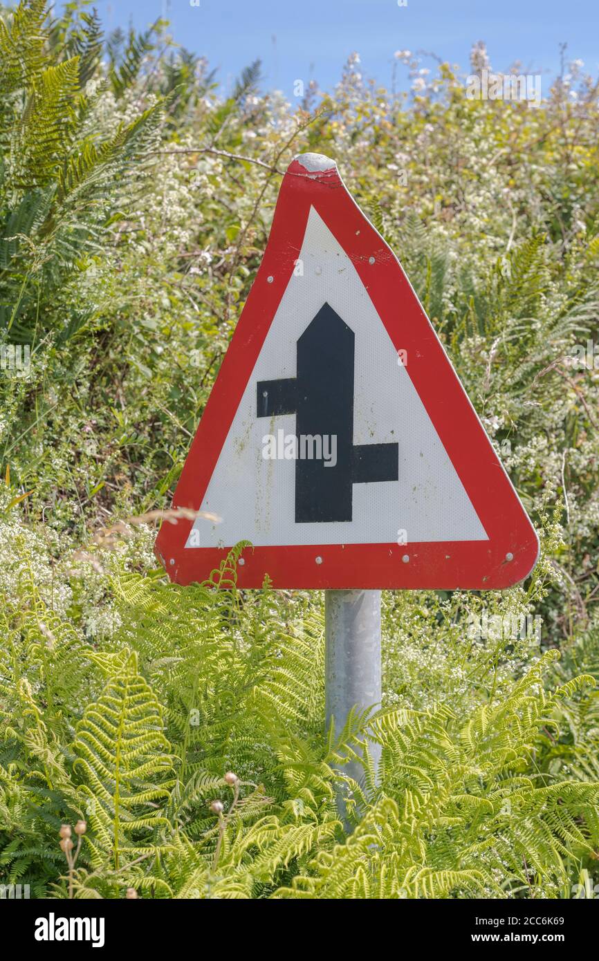Hedgerow rurale con il cartello stradale del Regno Unito che indica un bivio sfalsato davanti. Punto d'incontro metafora, traffico convergente, cartello di incrocio UK. Foto Stock