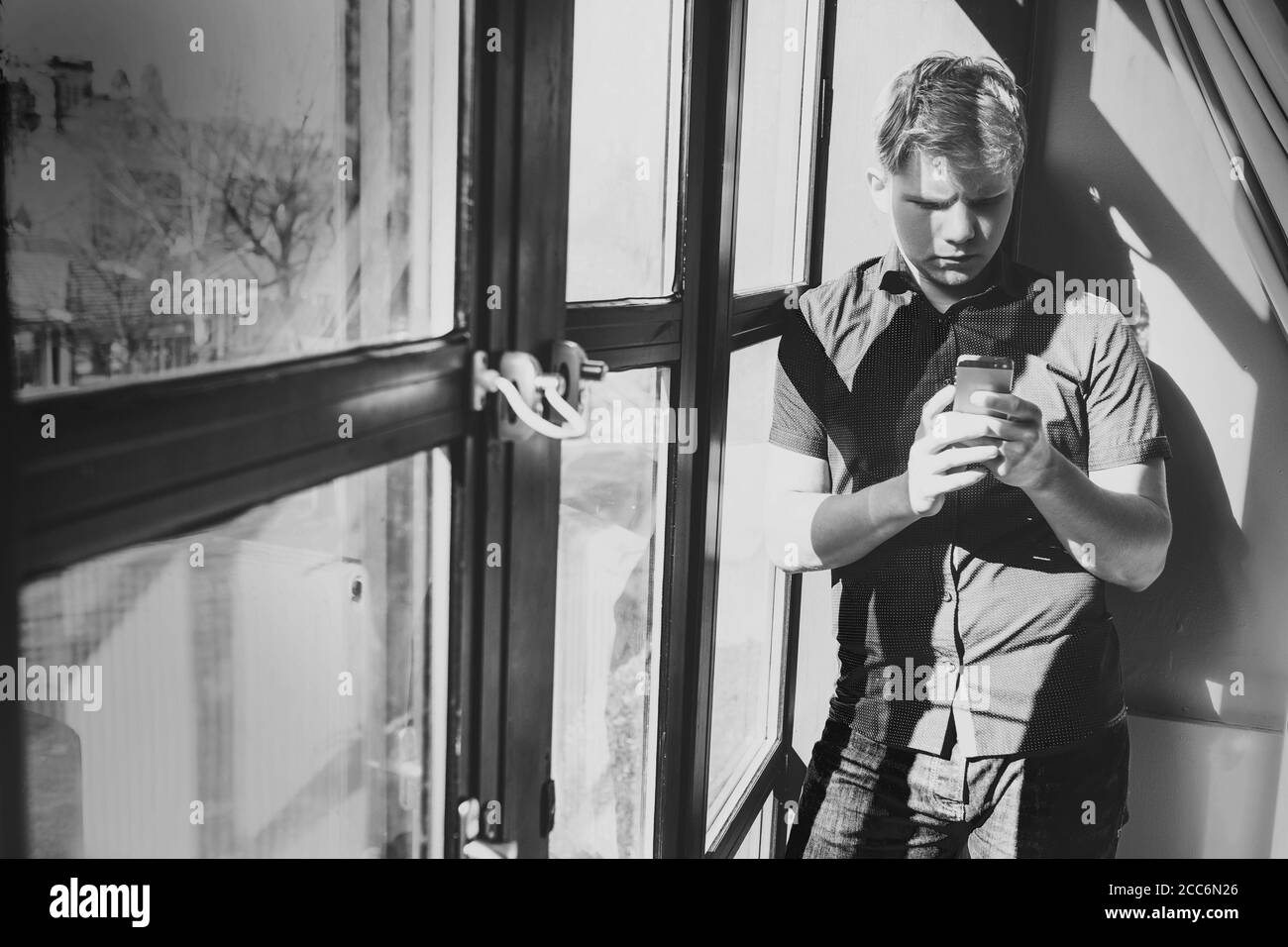 Giovane uomo con telefono cellulare stare vicino alla finestra dentro contrasto luce diurna Foto Stock
