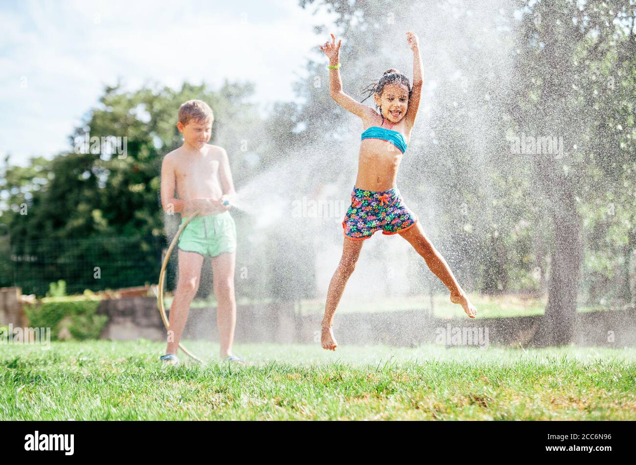 Attività calde dei giorni estivi: La bambina felice salta sotto l'acqua, quando il fratello la versa dal tubo del giardino Foto Stock