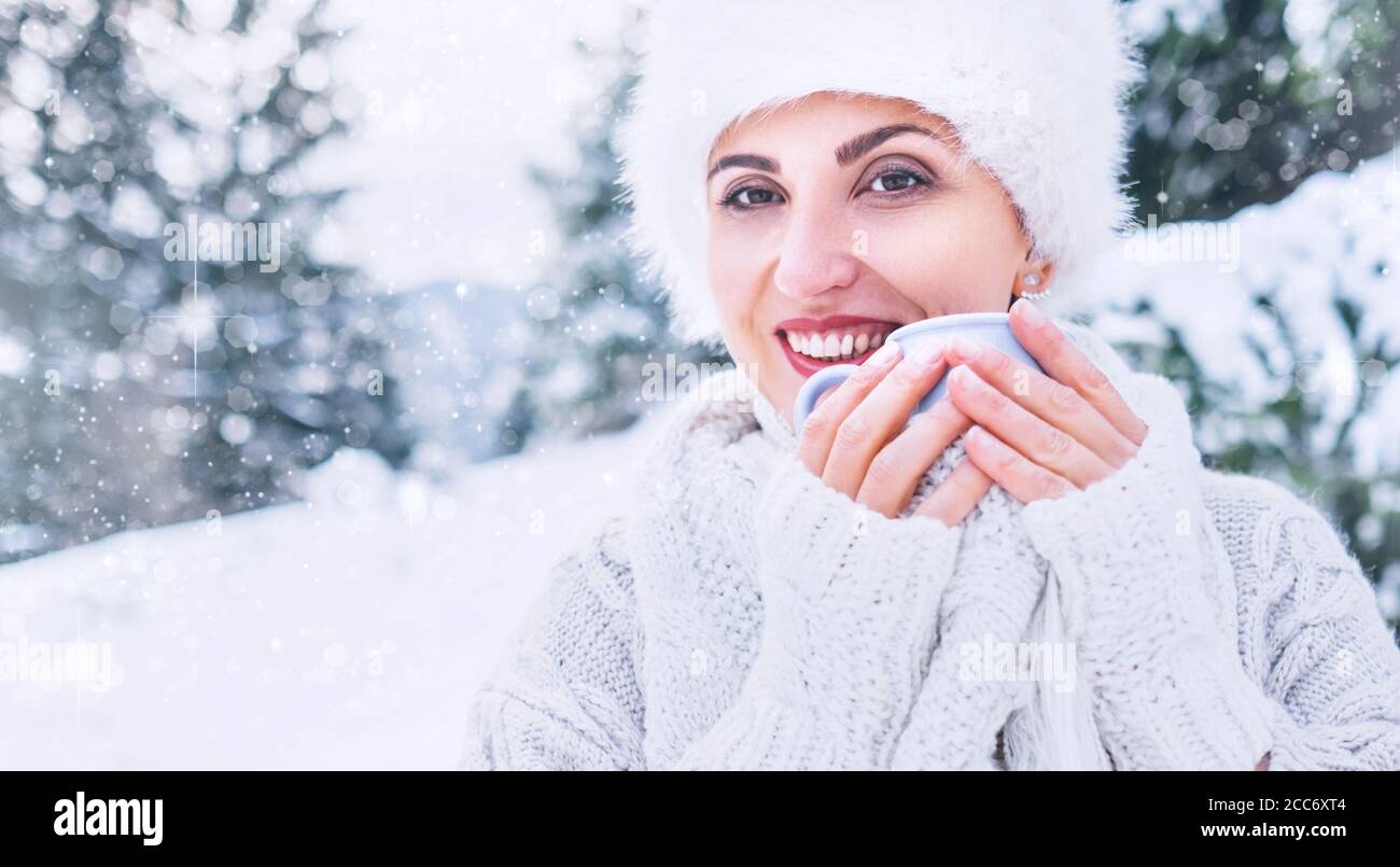 Donna sorridente in intimo maglieria e cappello di pelliccia con tazza di bevanda calda in inverno foresta medio shot. Concetto di moda invernale. Foto Stock