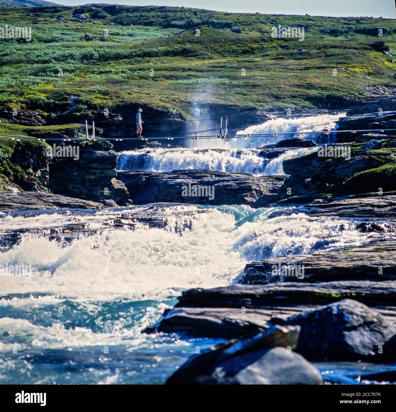 Uomo che attraversa un ponte sul fiume Staddajåkkå, in Svezia, sul lato orientale dei Monti Sulitjelma, in Nordland, Norvegia Foto Stock