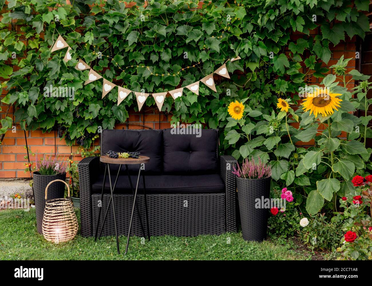 Sedia da giardino in polirotang moderna nera, tavolo laterale in filo metallico. Vitigni verdi piante che crescono su muro di mattoni rossi come elemento decorativo. Bamb Foto Stock