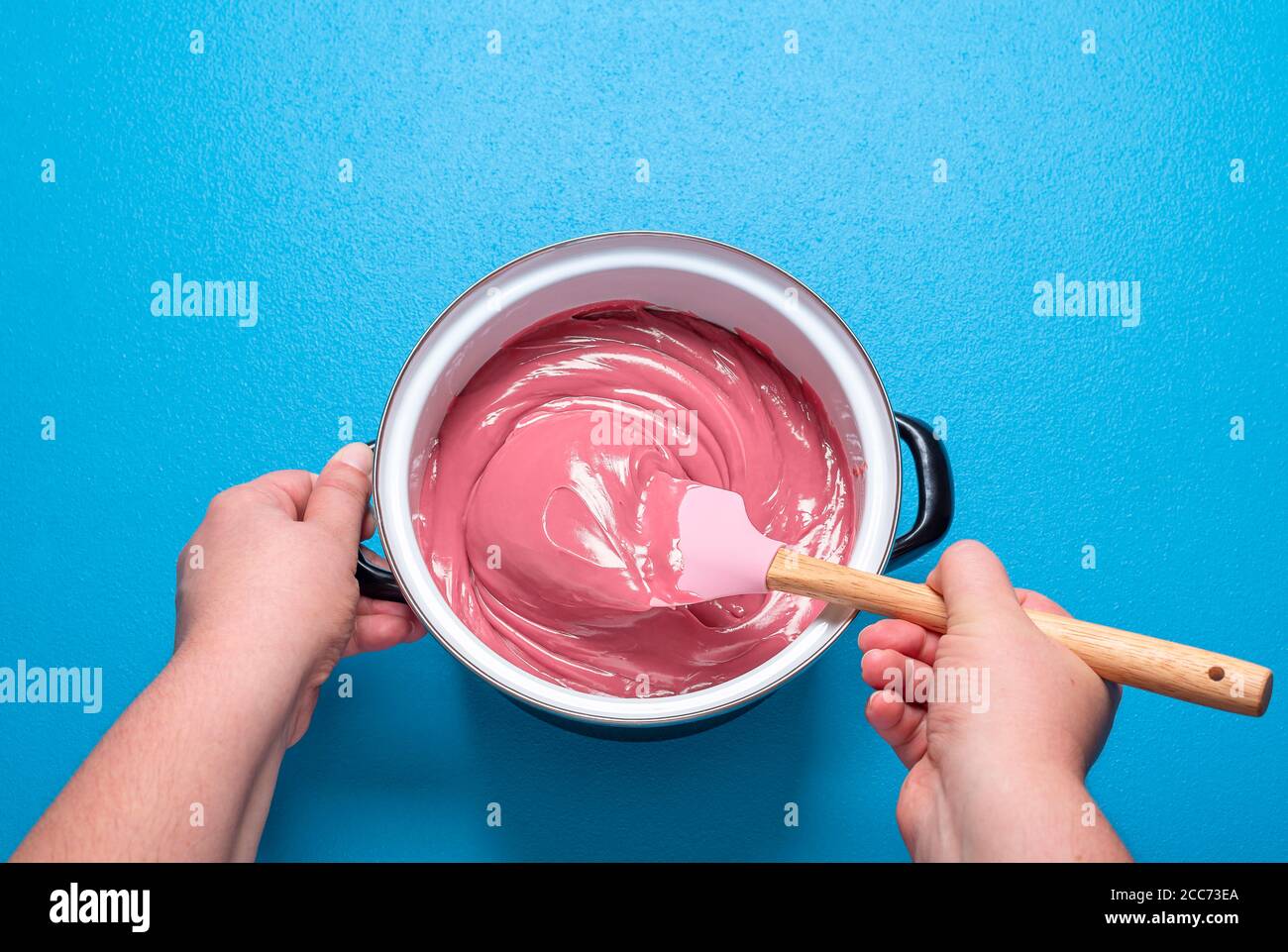 Donna che mescola lo smalto di cioccolato rubino in una pentola, su fondo blu. Vista dall'alto della turbolenza del cioccolato rosa fuso. Glassa rossa dolce. Cucinare a casa. Foto Stock