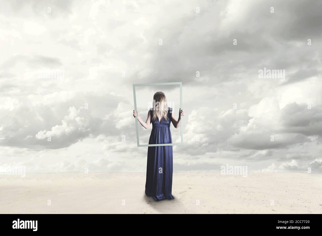 donna surreale con abito blu con faccia nascosta che tiene un cornice di una cornice vuota in un paesaggio marino Foto Stock