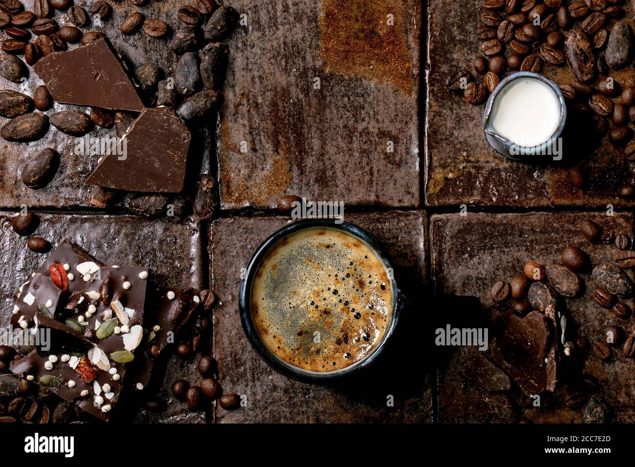Tazza di caffè espresso con caraffa di latte, cioccolato fondente fatto a mano, caffè e chicchi di cacao intorno a piastrelle di ceramica scura come sfondo. Posa piatta, copia s Foto Stock