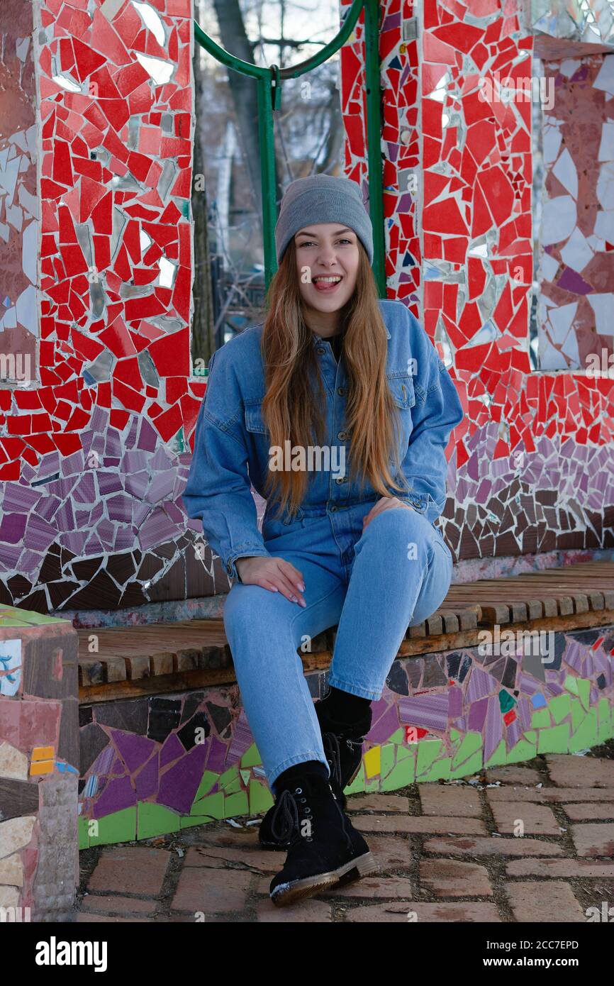 donna in tuta in denim blu e cappello grigio siede su una panchina vicino ad una parete di glass.the colorato donna mostra la sua lingua e sorrisi Foto Stock