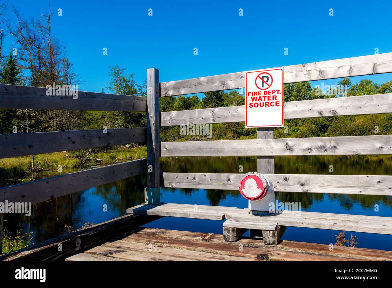 Un segno che dice fonte di acqua del reparto di fuoco su una recinzione di legno. Nessun simbolo di parcheggio sopra il testo. Collegamento del tubo antincendio sotto. Stagno dietro recinzione, alberi in backg Foto Stock