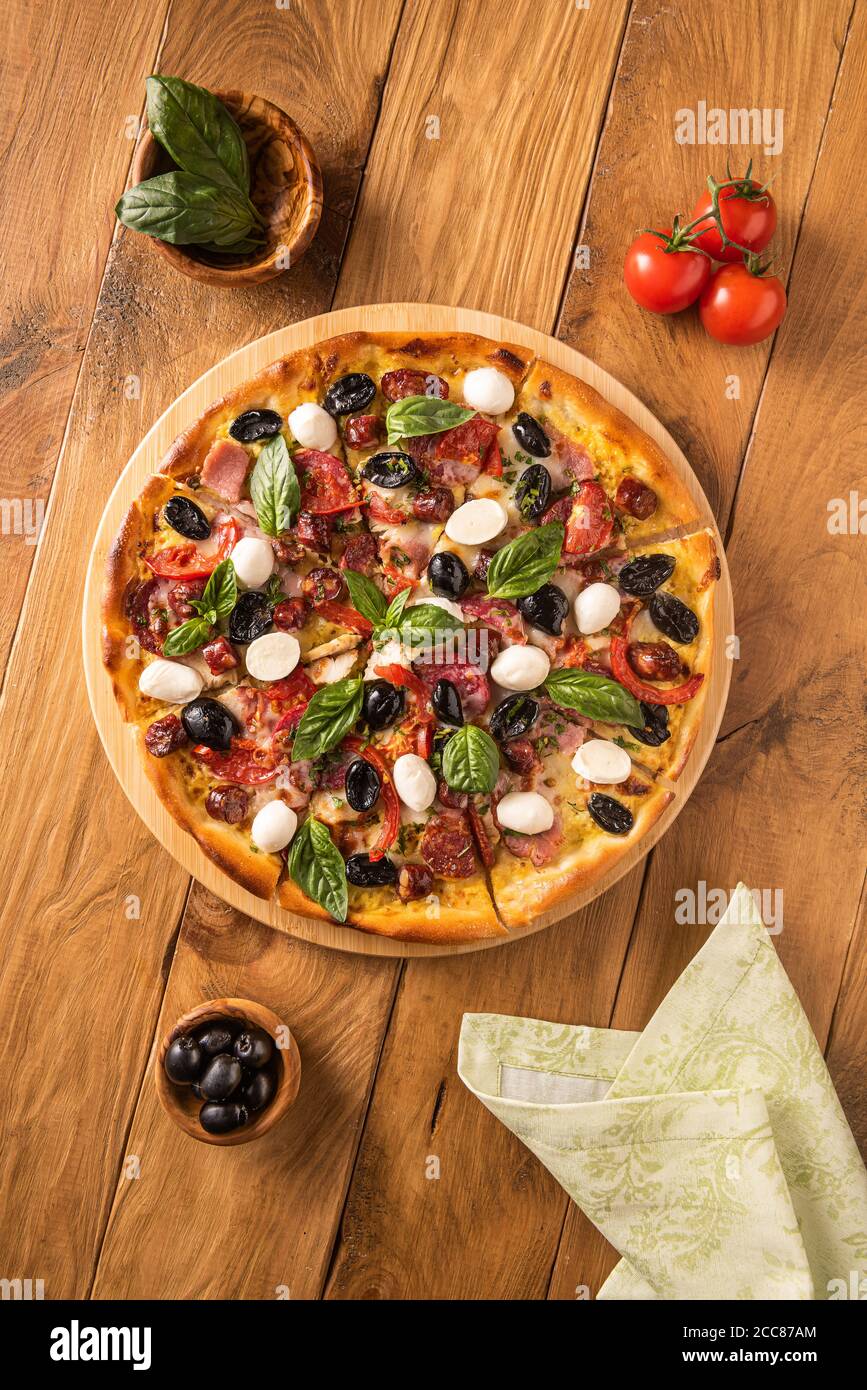 Pizza italiana con prosciutto, salame, olive nere, mozzarella, pomodori rossi e foglie di basilico su fondo ligneo e pochi ingredienti in ciotola di legno Foto Stock