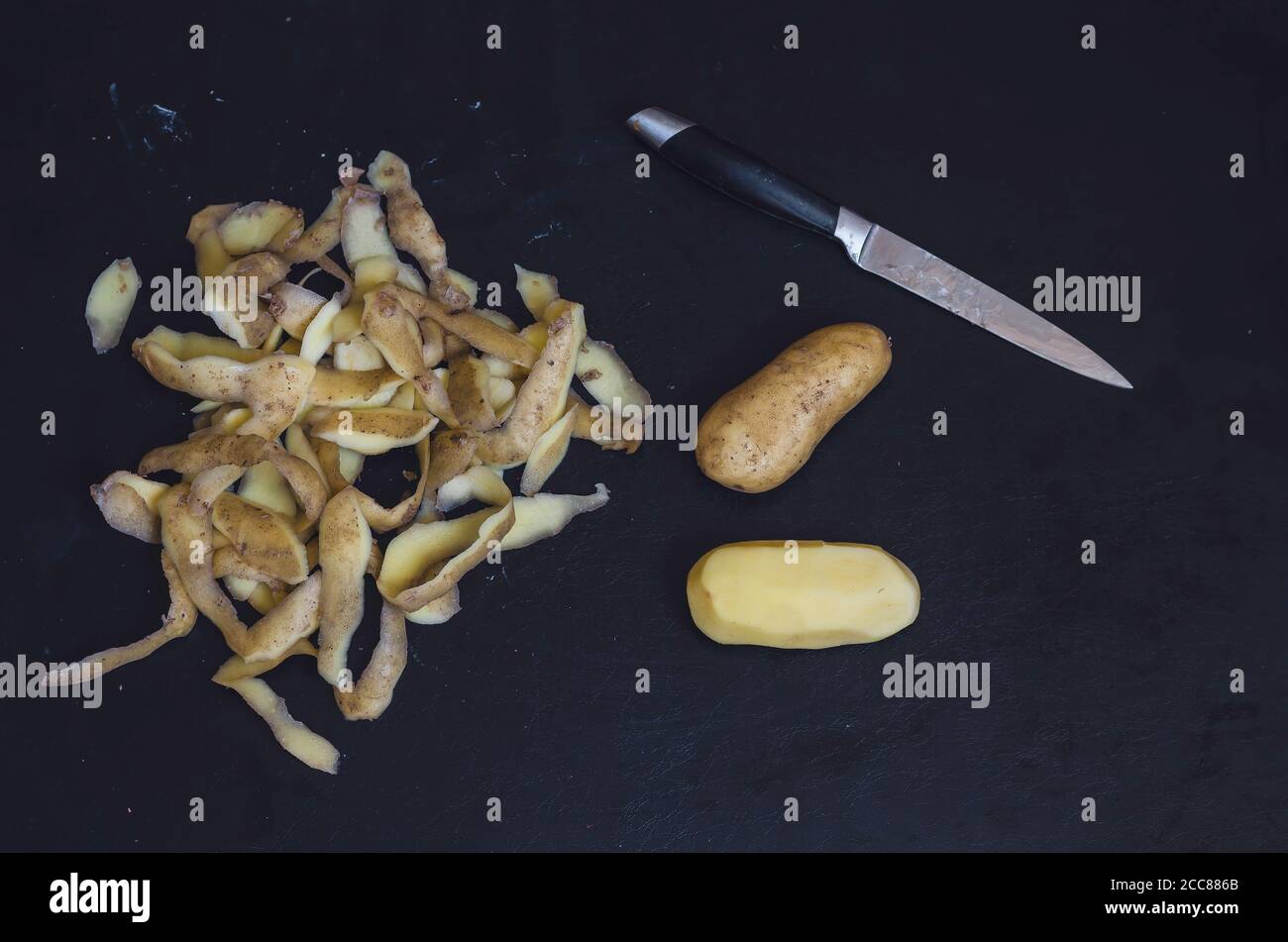 Due diverse patate crude, pulizia e un coltello sul tavolo nero. Radici in e senza buccia accanto allo spreco di cibo. Cucina di cibo. Messa a fuoco selettiva. Foto Stock