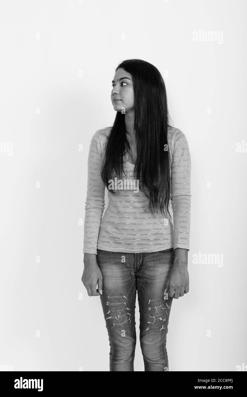 Vista di profilo di giovane donna indiana in piedi mentre pensando con corpo rivolta verso la telecamera contro uno sfondo bianco Foto Stock