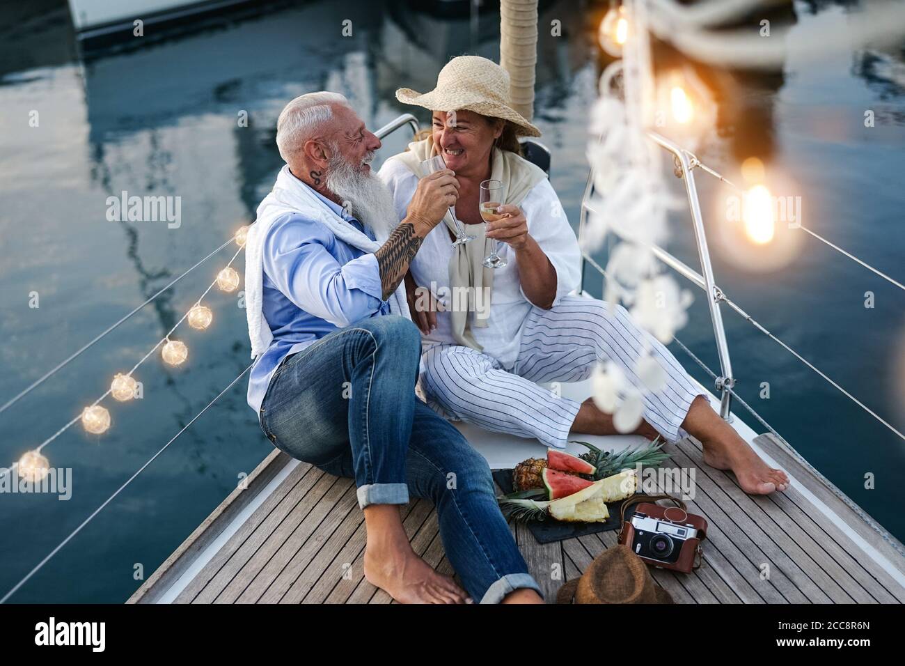 Coppia senior che brindano champagne e mangiano frutta in vacanza in barca a vela - persone anziane felici che si divertono a celebrare l'anniversario di matrimonio gita in barca Foto Stock