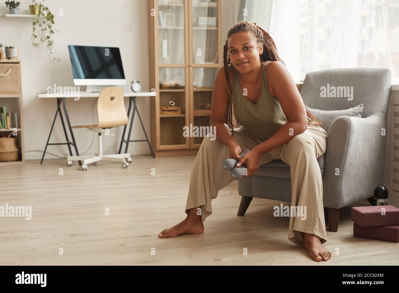 Ritratto di giovane donna africana seduta su poltrona con manubri e guardando la macchina fotografica che riposa dopo l'addestramento Foto Stock