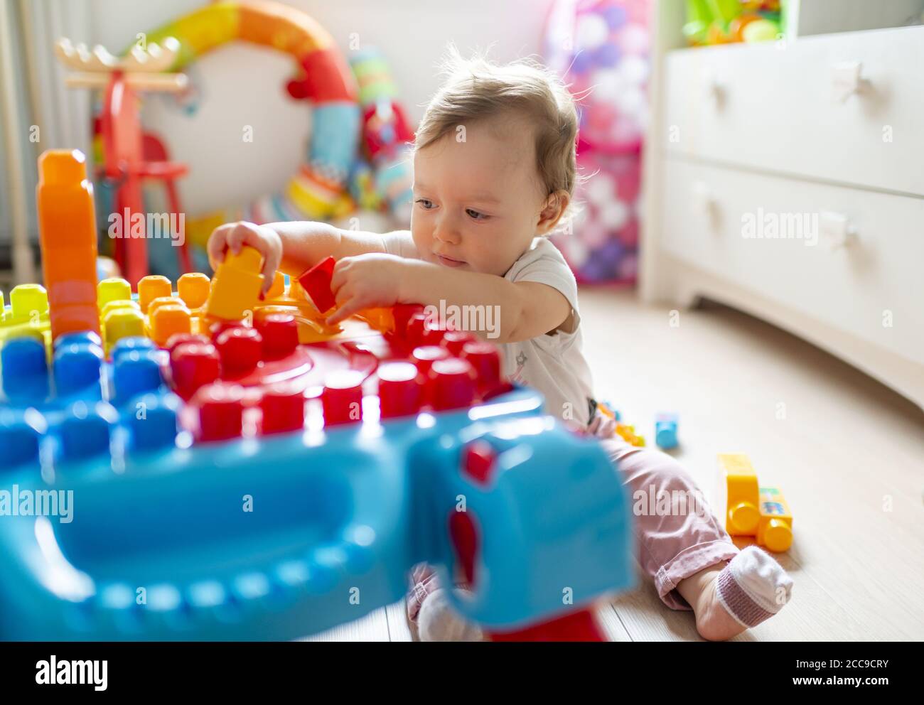 Bambina (16 mesi) seduta sul pavimento e giocando nella sua camera da letto con Colorati blocchi di plastica Mega Bloks® vicino al suo petto di legno bianco di cassetti Foto Stock