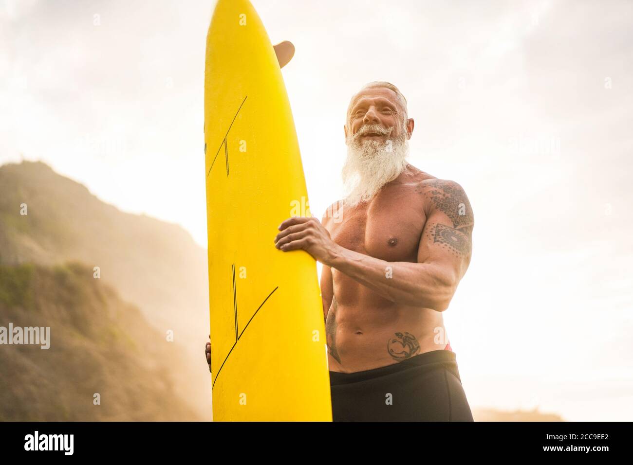 Tattooed surfer senior tenendo surf board sulla spiaggia al tramonto - ragazzo felice vecchio divertirsi facendo sport estremo - gioioso anziano concetto - Focus on hi Foto Stock