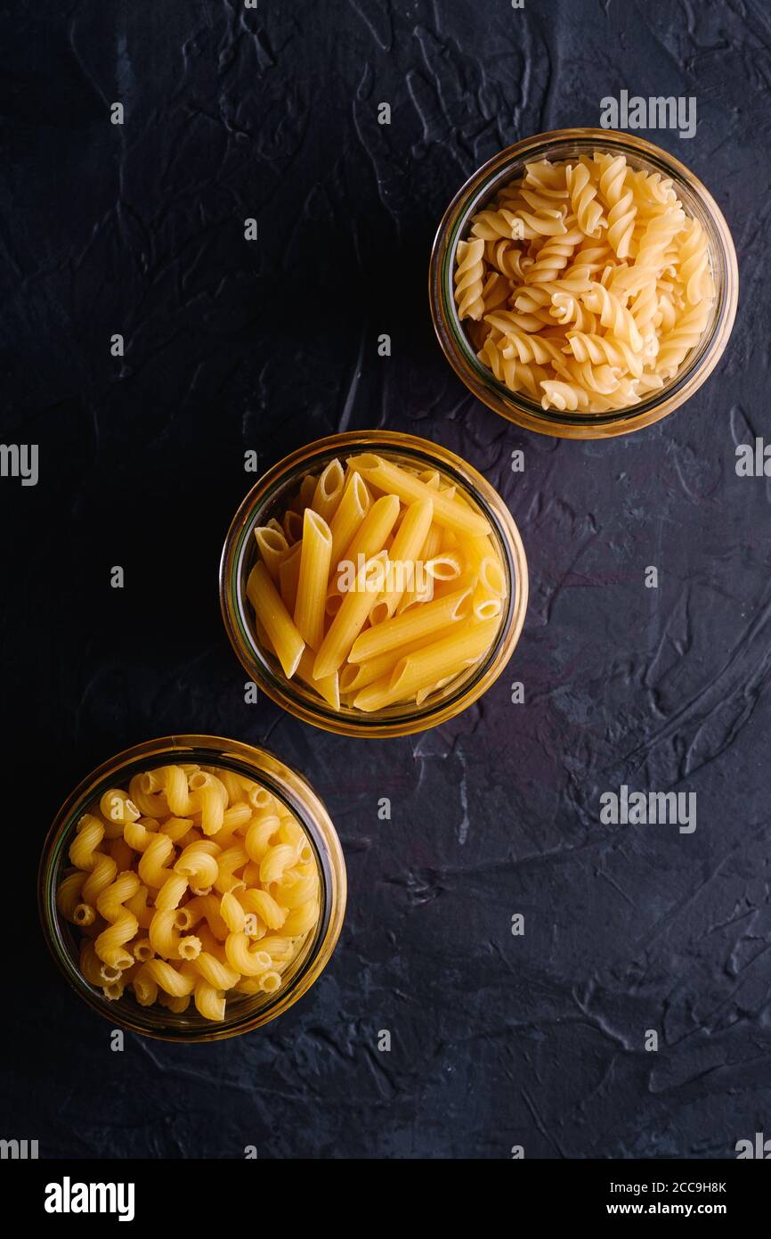 Tre vasetti di vetro in fila con una varietà di pasta di grano dorato non cotta su sfondo nero scuro testurizzato, vista dall'alto Foto Stock