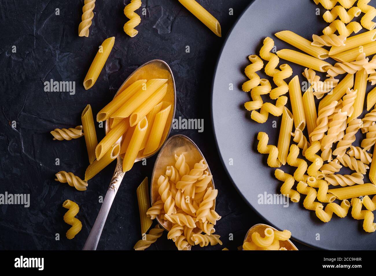 Tre cucchiai di posate e piatto con una varietà di pasta di grano dorato non cotta su sfondo nero scuro testurizzato, macro vista dall'alto Foto Stock