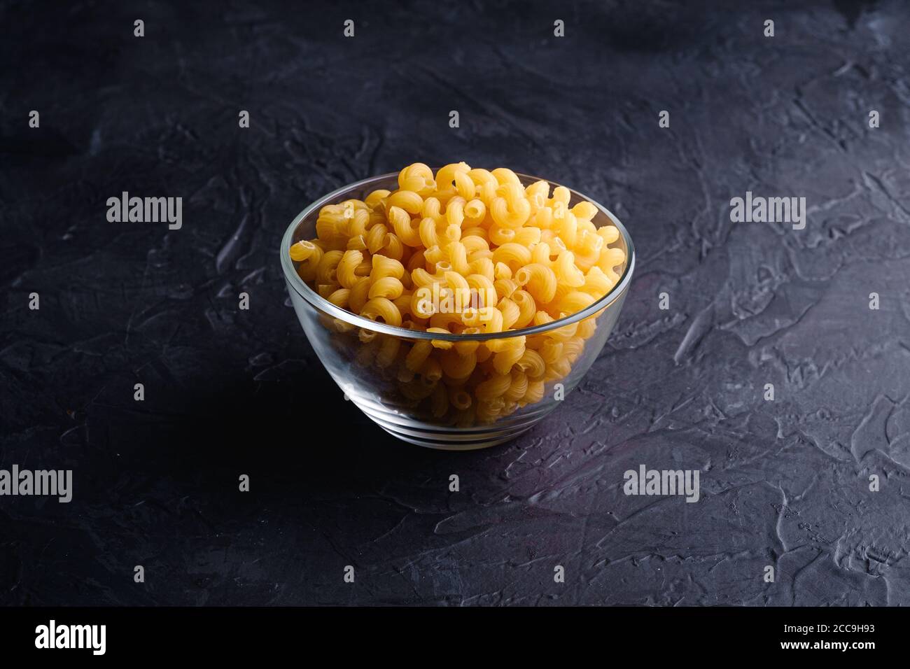 Ciotola in vetro con cavatappi pasta riccia di grano dorato crudo su fondo nero scuro testurizzato, vista angolare Foto Stock