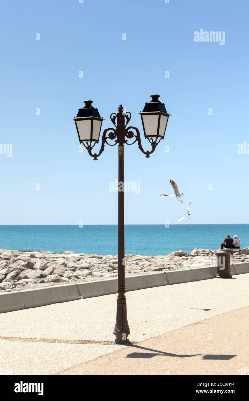Lanterna su una passerella sul Mar mediterraneo, due persone non identificabili seduti sulle rocce, due gabbiani che volano Foto Stock