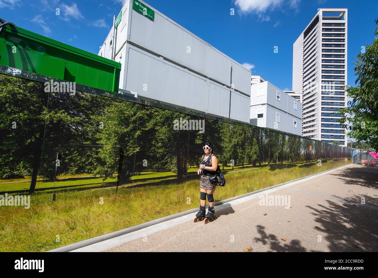 Un rollerblader passa un'accatastamento che mostra una scena di verde bosco, al di fuori di un cantiere nel nord di Londra, mentre la gente apprezza il caldo dopo le recenti forti precipitazioni piovose. Foto Stock