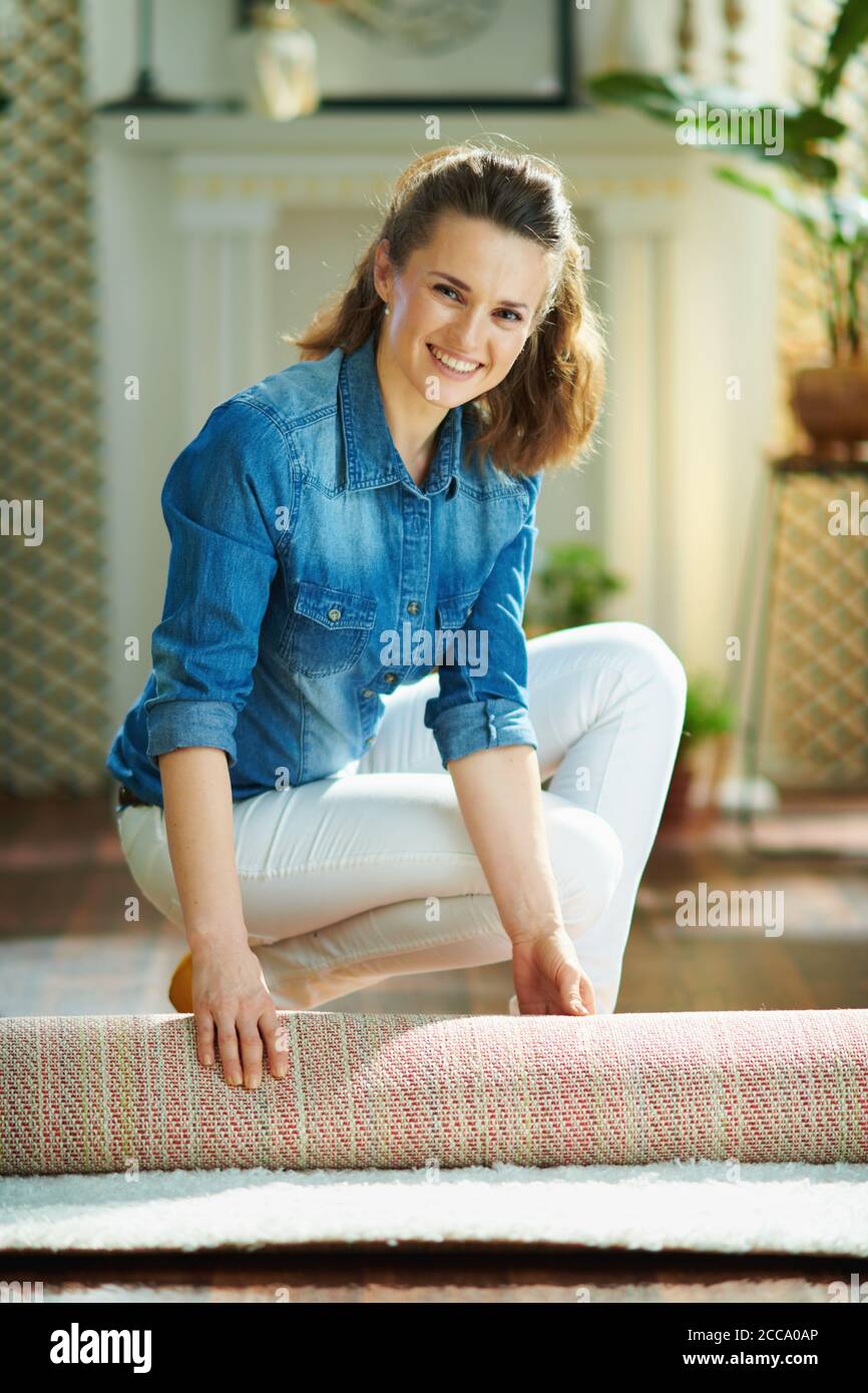 sorridente donna moderna di 40 anni in jeans camicia e pantaloni bianchi  nel moderno soggiorno in giornata di sole moquette pieghevole in un rotolo  Foto stock - Alamy