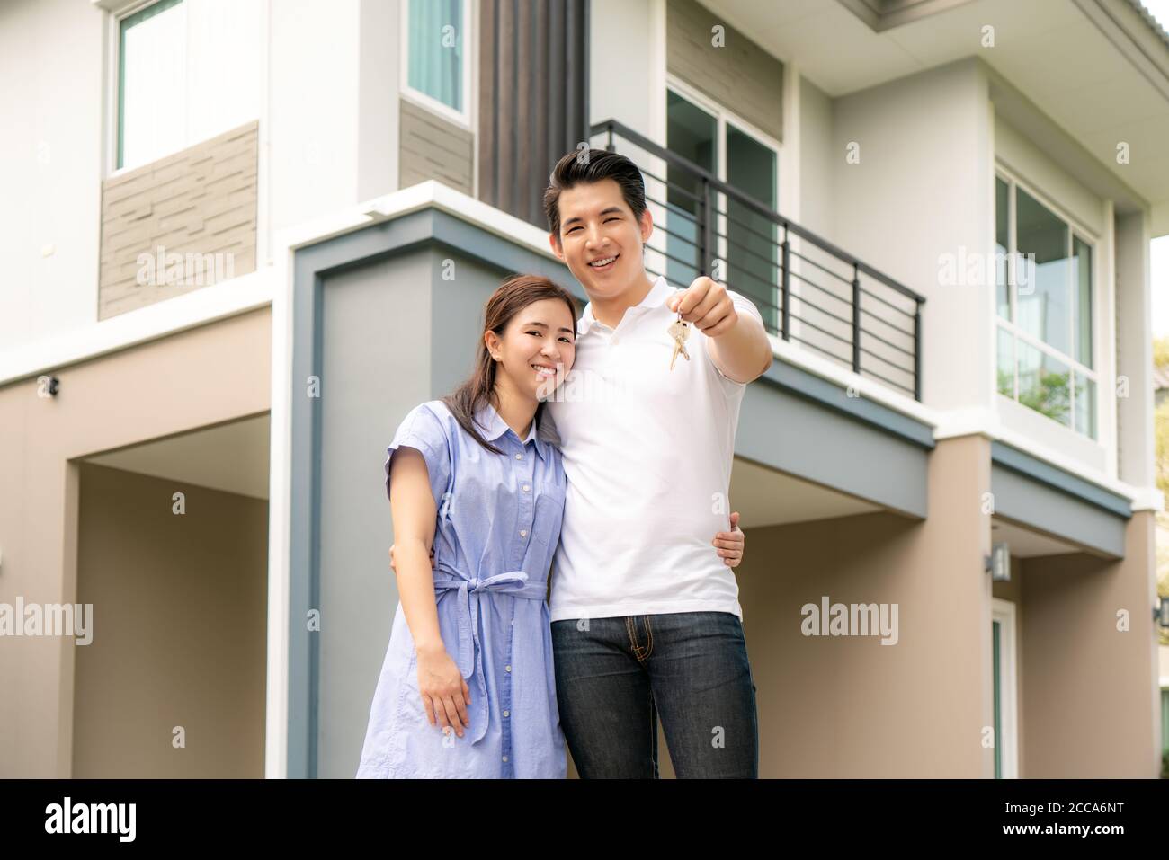 Ritratto di giovane coppia asiatica in piedi e abbracciando insieme e tenendo la chiave della casa che guarda felice davanti alla loro nuova casa per iniziare la nuova vita. Famiglia Foto Stock