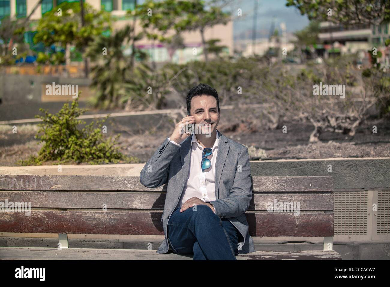 Adulto sorridente ben vestito imprenditore maschile seduto su panca di legno e discutere di notizie durante la telefonata mentre si è in pausa e. riposando sulla strada in Foto Stock