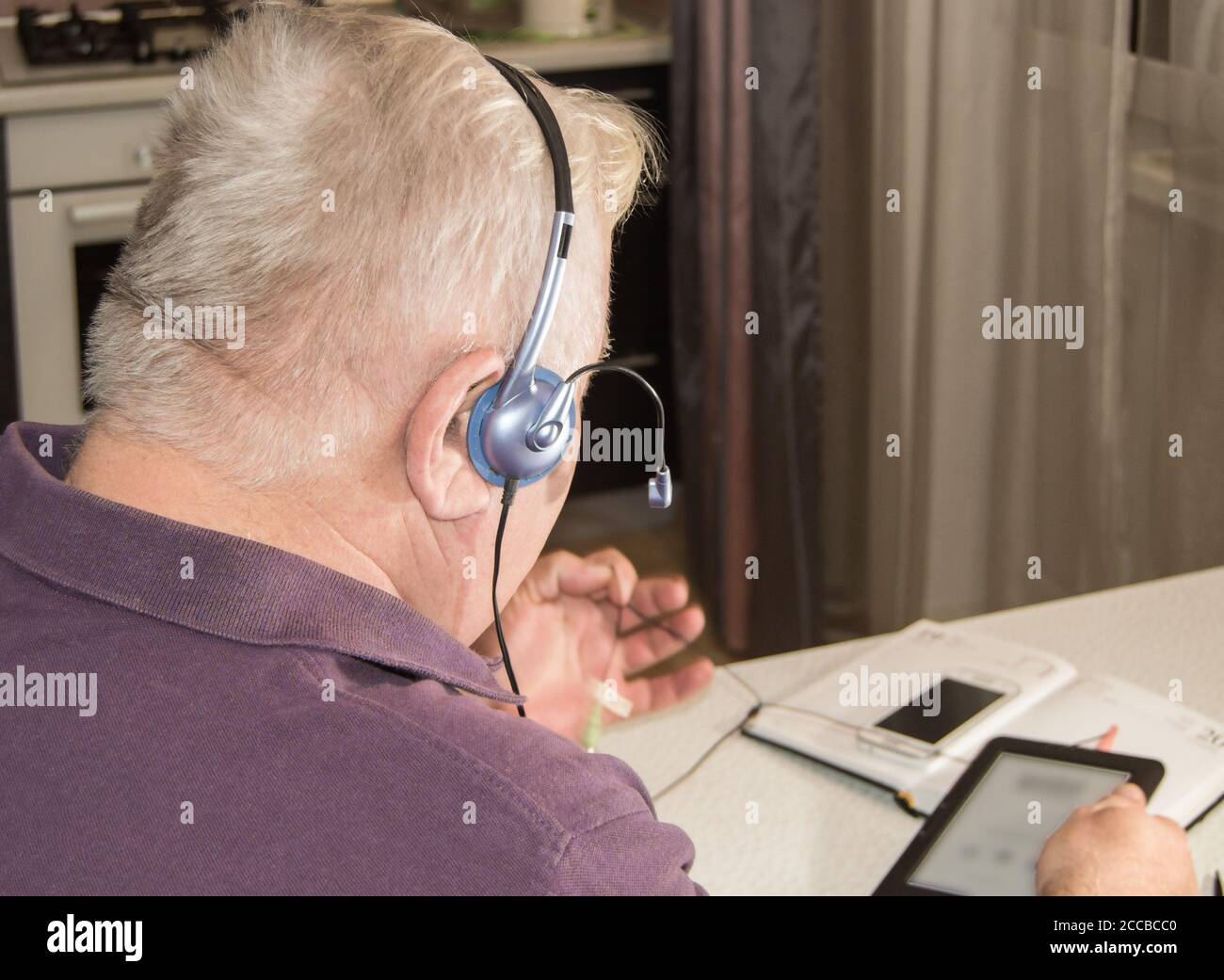 Un insegnante anziano dai capelli bianchi in un seminario online con un tablet, una fotocamera, un telefono e una cuffia, un webinar presso un'università di distanza, una scuola, un tutor, un tel Foto Stock