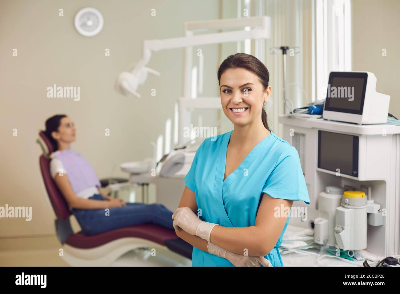 Dentista sorridente in piedi e guardando la fotocamera con il paziente seduto sullo sfondo Foto Stock