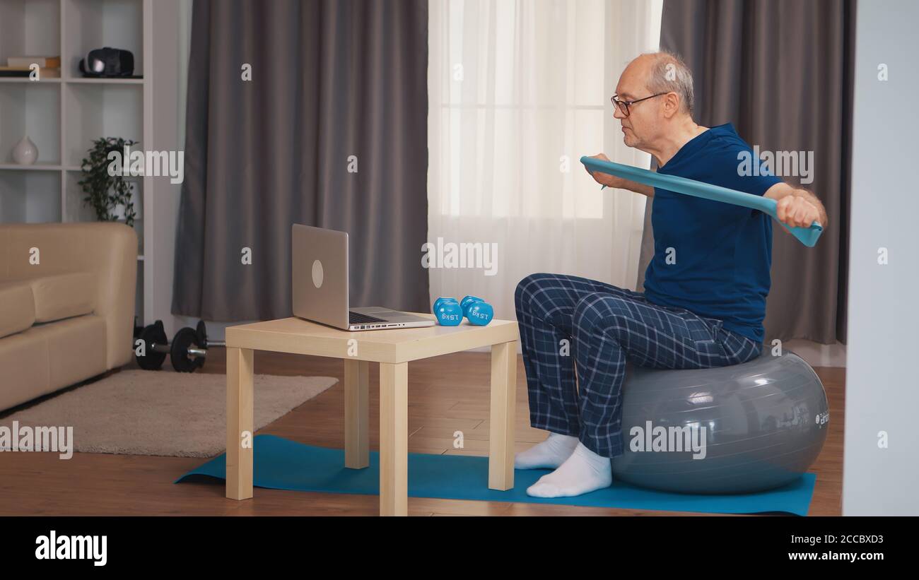 L'uomo anziano sull'addestramento della sfera di equilibrio con la banda di resistenza che guarda il programma in linea di idoneità. Anziano pensionato sano addestrando sport di salute a casa, esercitandosi attività di idoneità all'età anziana Foto Stock